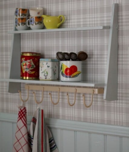 Vitmålad liten hylla med plåtburkar, tekoppar och andra saker uppsatt på rutig köksvägg.