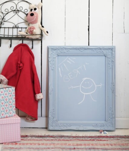 Liten ljus tavelram målad i samma färg till en griffeltavla, innanför ramen står det skrivet med skolkrita "Bertil" och bredvid är en glad gubbe.