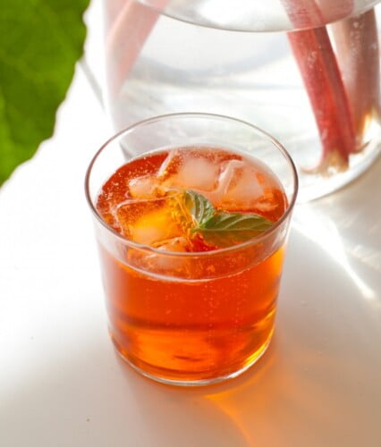 Guldfärgad drink med mycket is i glaset på sommarbord intill vas med rabarber.