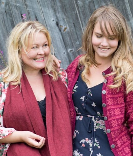 Erica och Clara skrattar, klädda i vinröda toner utanför grå brädlada.