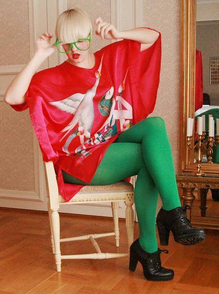 Clara i blont rakt hår, röd överdel och knallgröna strumpbyxor och gröna glasögonbågar sitter på en stol framför guldspegel.