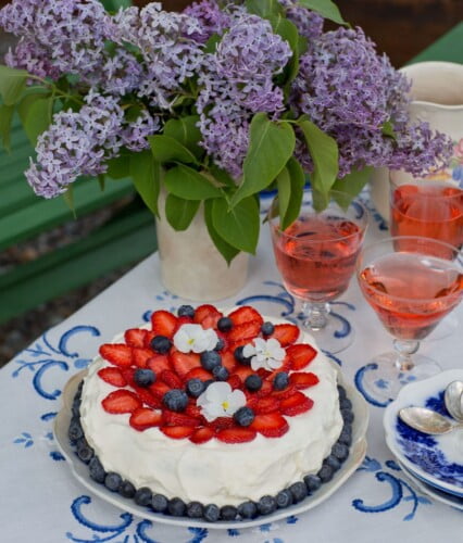 härlig gräddtårta med skivade jordgubbar och blåbär under bukett av syrener på dukat sommarbord.