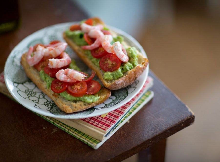 Lunchsmörgås i två delar med skaldjur, avokadoröra och tomat på. Ligger på tallrik som står på en träpall.