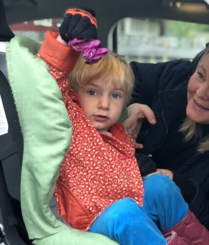 Folke sitter i bilen med ytterkläder på och håller fram en bit rödkål som han ska ha med till förskolan. Erica hjälper honom att fästa bältet i barnstolen. Ur En Underbar Pod.