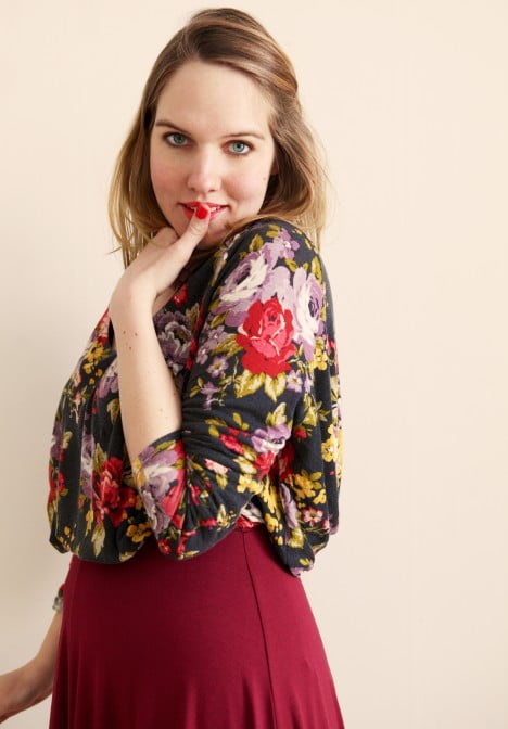 Clara i blommig blus och vinröd långkjol tittar in i kameran med försiktigt leende.