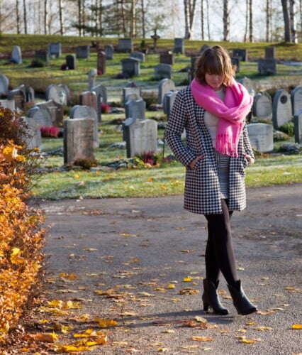 Clara går i höstsol på kyrkogård i rutig kappa och rosa halsduk.