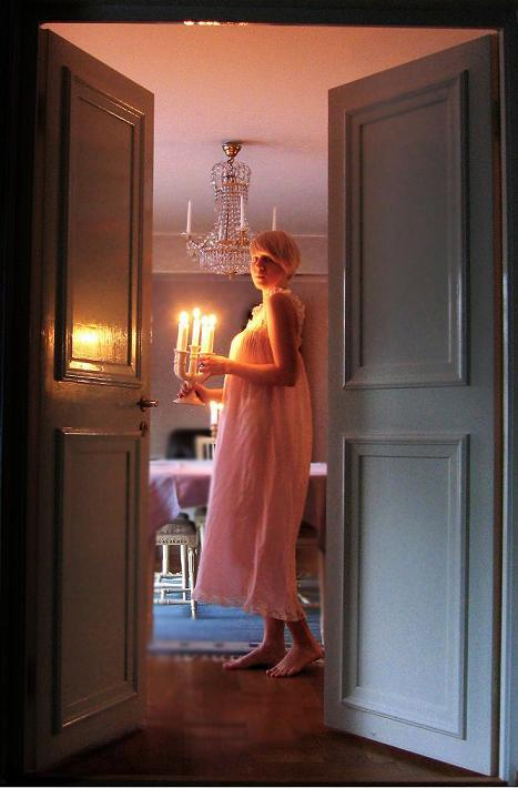 Clara i vitt långt nattlinne, bär en kandelaber med levande ljus genom ett mörkt rum.