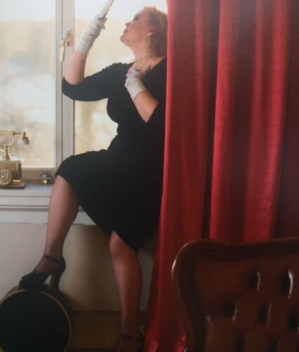 Erica sitter i fönster i mörk sammetsklänning så valkar syns i midjan. Ur En Underbar Pod. Tjica.