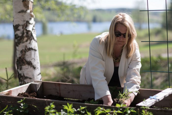 Erica planterar blommor i låda iklädd ljus kappa och solglasögon. Ur En Underbar Pod.