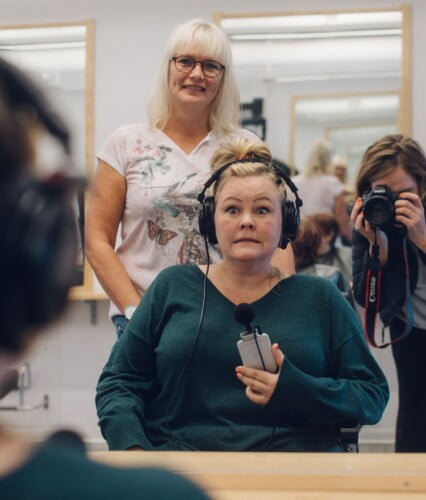 Erica sitter och stirrar oroligt in i spegeln hos frisören Marlene. Clara fotar så hon också syns i spegeln. Ur En Underbar Pod.
